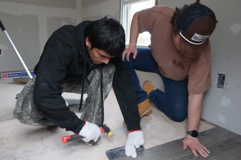 Two men work together on installing flooring in a room under renovation. One kneels and uses a hammer and flooring tool while the other braces the plank in place, watching closely.